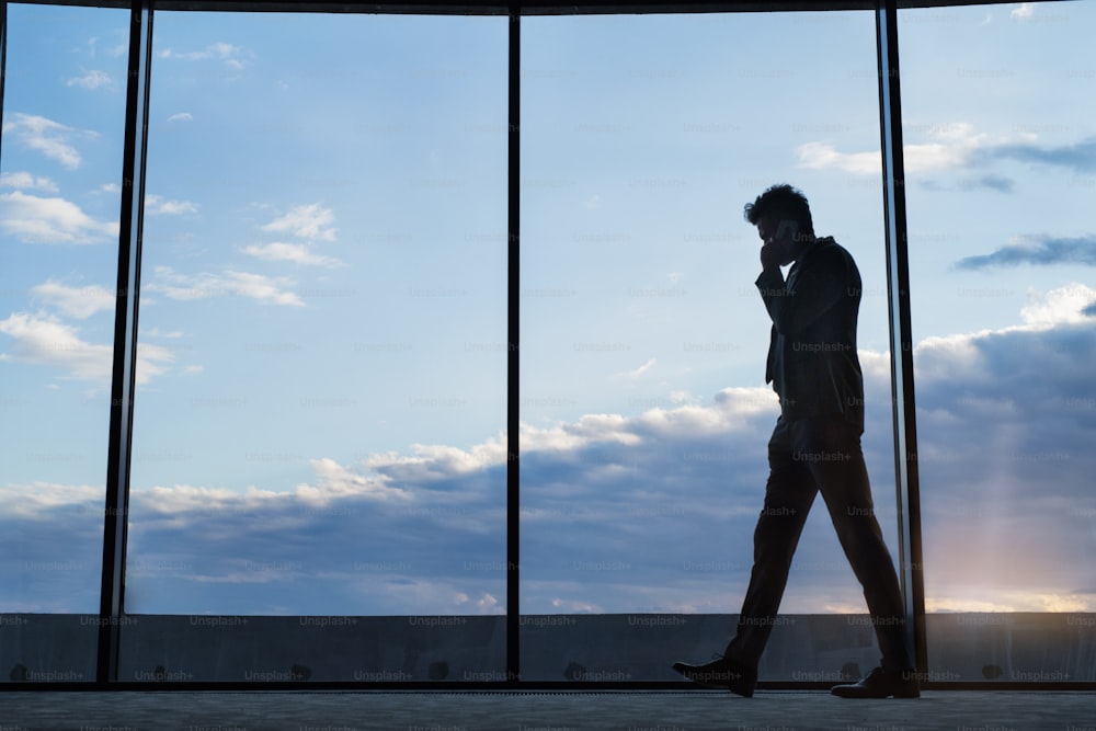 Homme d’affaires mature dans un hôtel avec un smartphone. Homme faisant un appel téléphonique, marchant le long de grandes fenêtres. Homme d’affaires mature dans un hôtel avec un smartphone. Homme faisant un appel téléphonique, marchant le long de grandes fenêtres.