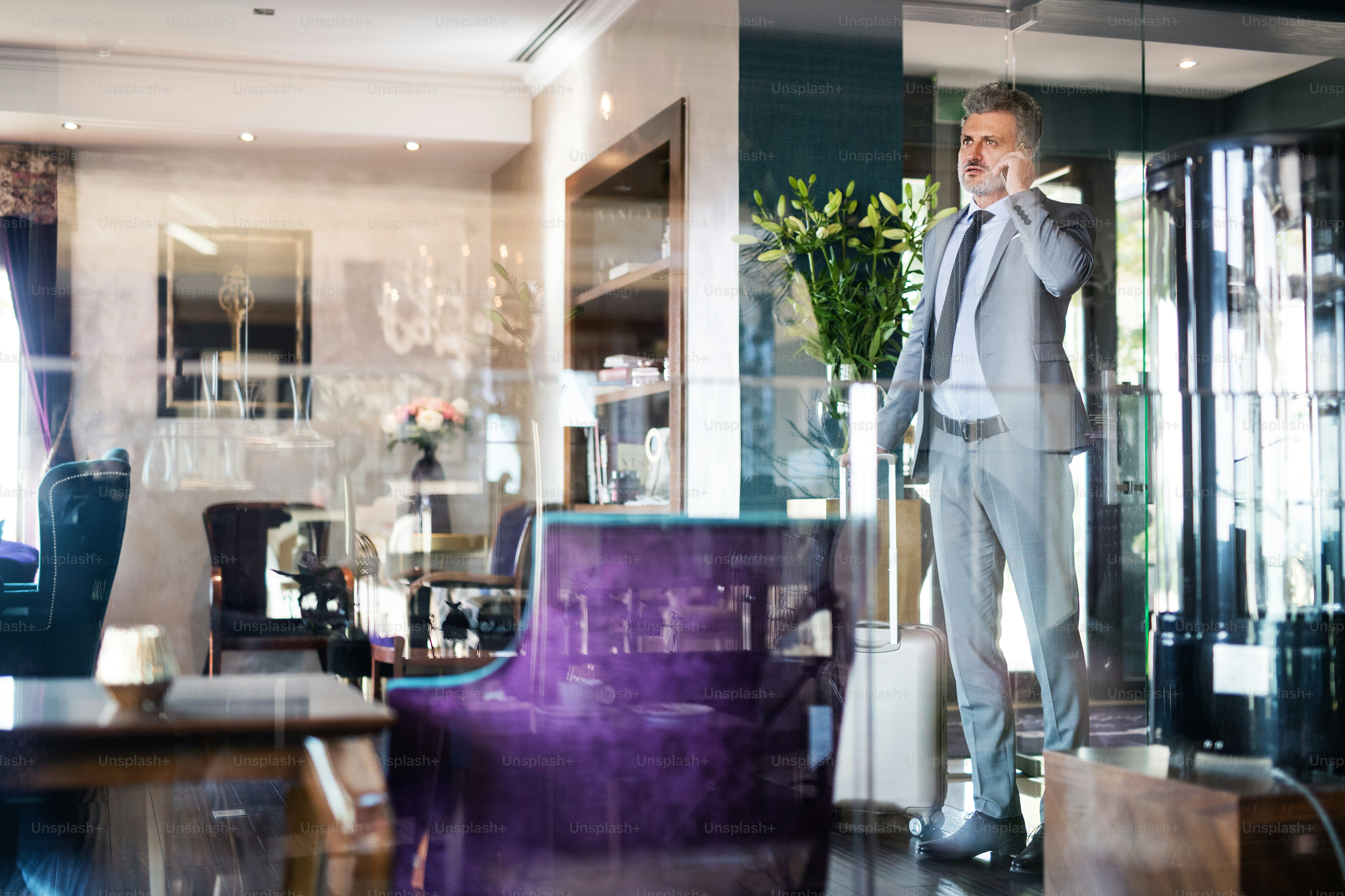 Handsome mature businessman with smartphone in a hotel lounge, making a phone call. Shot through glass.