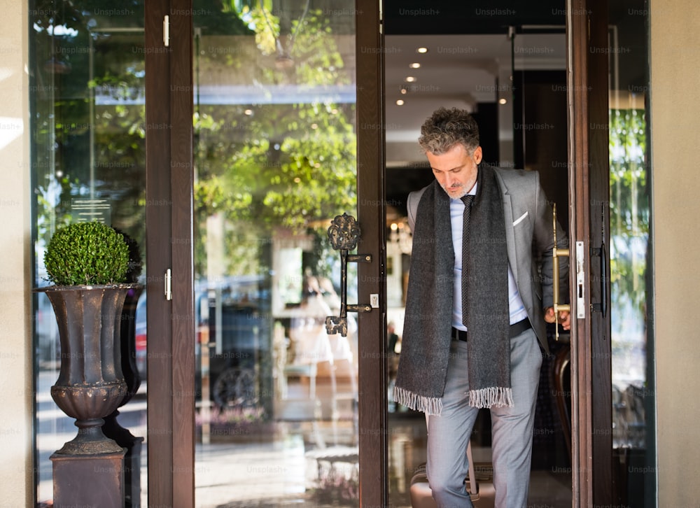 Mature businessman leaving hotel with luggage. Man walking out through the hotel front door.