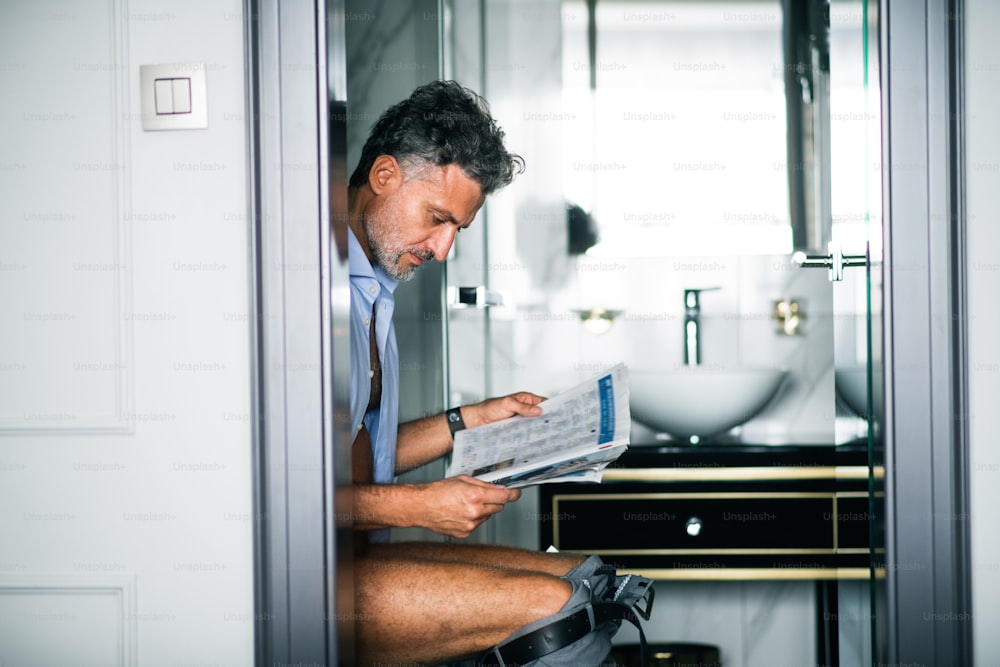 Uomo d'affari maturo in un bagno della camera d'albergo. Bel uomo seduto sul water, leggendo i giornali.