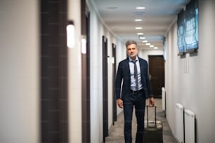 Uomo d'affari maturo che cammina con i bagagli in un corridoio dell'hotel. Uomo bello che tira la valigia.