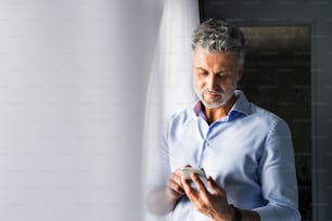 Uomo d'affari maturo con smartphone in una stanza d'albergo. Bel uomo in piedi alla finestra, messaggi. Primo piano.