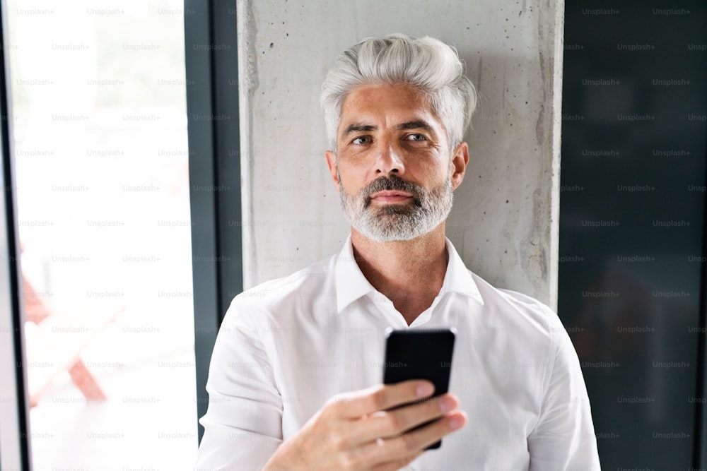 Hombre de negocios maduro con camisa blanca en la oficina parado en la pared de concreto, sosteniendo un teléfono inteligente, enviando mensajes de texto.