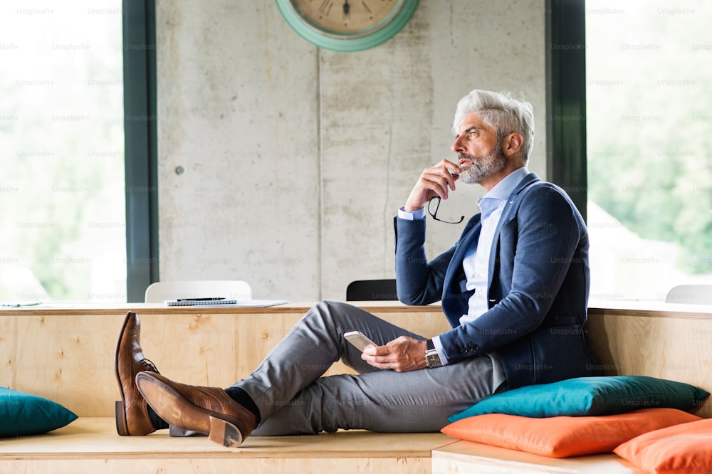 Homme d’affaires mûr et réfléchi avec smartphone au bureau, assis avec les jambes sur le bureau.