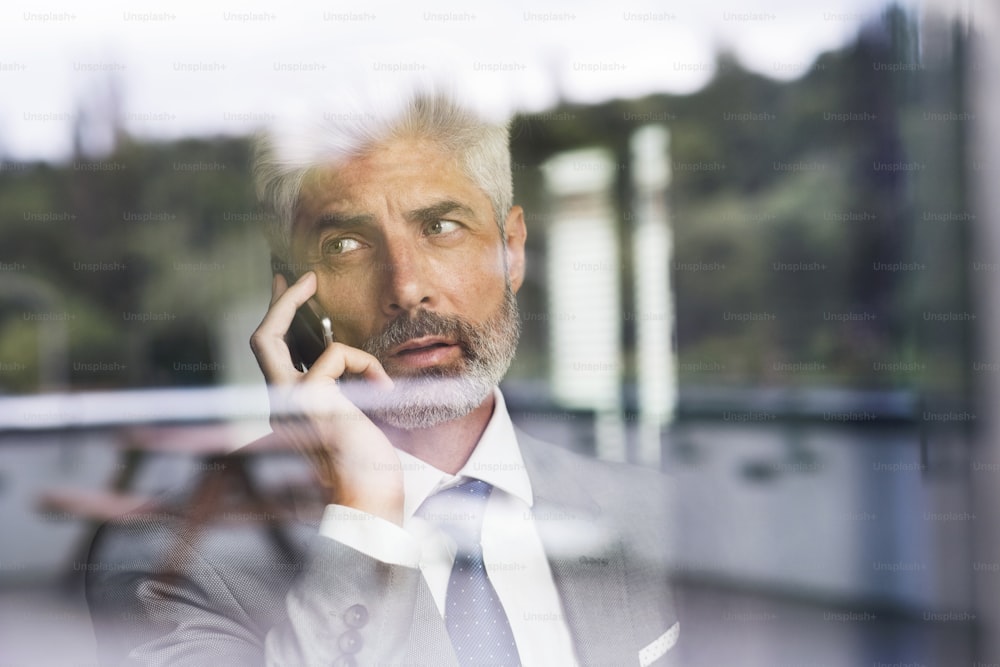 Homme d’affaires mature en costume gris dans le bureau debout à la fenêtre tenant un smartphone en train de passer un appel téléphonique. Tiré à travers le verre. Gros plan.