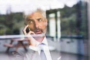 Maturo uomo d'affari in abito grigio in ufficio in piedi alla finestra con lo smartphone che fa telefonata. Girato attraverso il vetro. Primo piano.