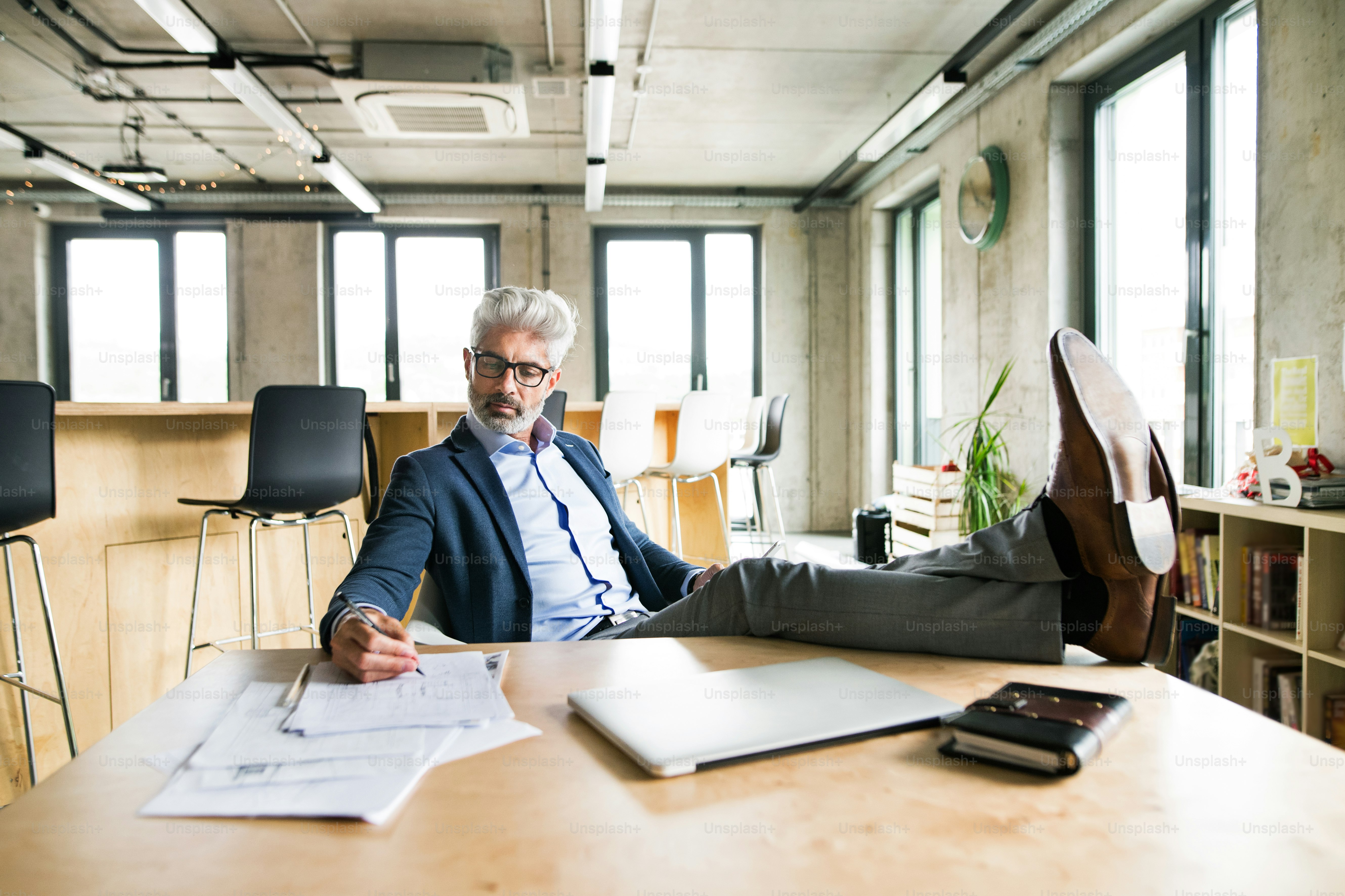 Nachdenklicher reifer Geschäftsmann mit grauen Haaren im Büro, der mit den Beinen auf dem Schreibtisch sitzt und arbeitet.