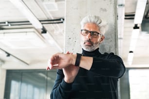 Handsome mature businessman with gray hair in black turtleneck and eyeglasses in the office. Man checking time on his watch.