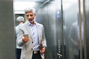 Maturo uomo d'affari in piedi nell'ascensore con in mano lo smartphone, messaggi.