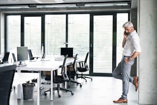 Mature businessman in white shirt in the office standing, holding smartphone and making phone call.