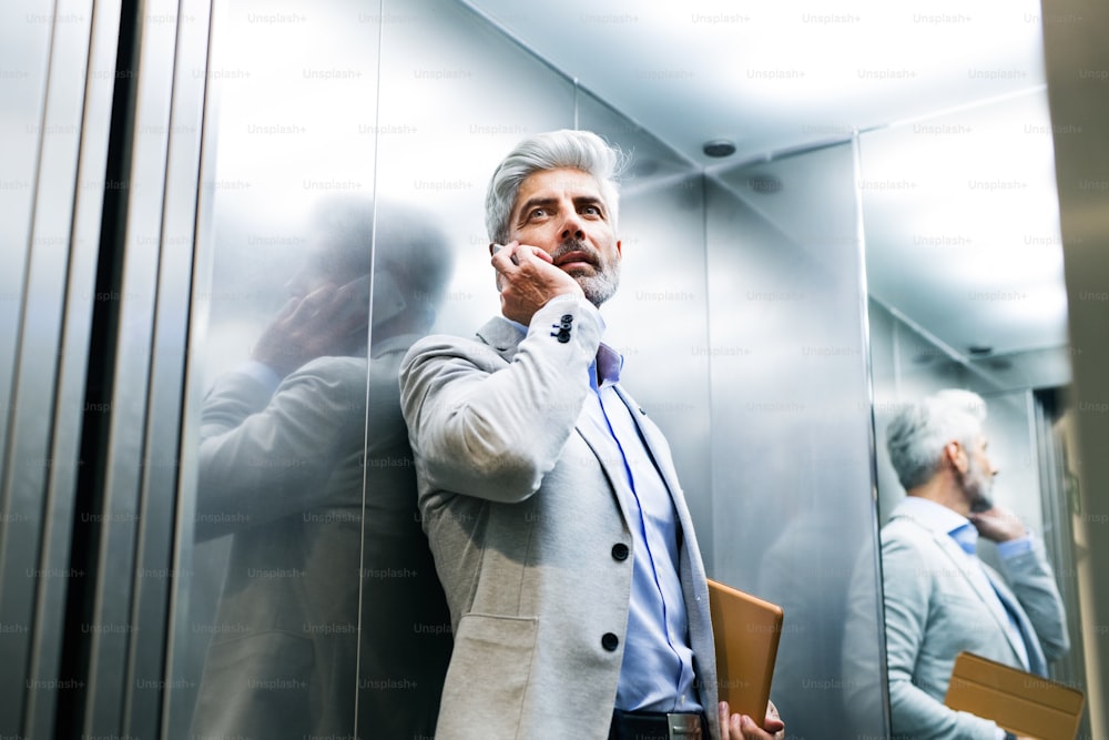 Mature businessman standing in the elevator holding smartphone making phone call.