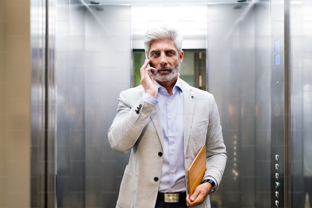Hombre de negocios maduro en la oficina de pie en el ascensor sosteniendo un teléfono inteligente haciendo una llamada telefónica.