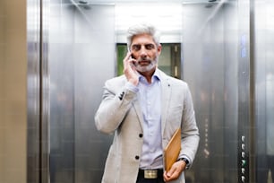 エレベーターに立って電話をかけるスマートフォンを持つオフィスの成熟したビジネスマン。