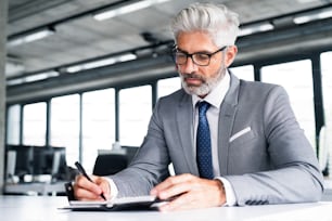Hombre de negocios maduro con traje gris sentado en el escritorio de la oficina, escribiendo algo en su organizador personal.