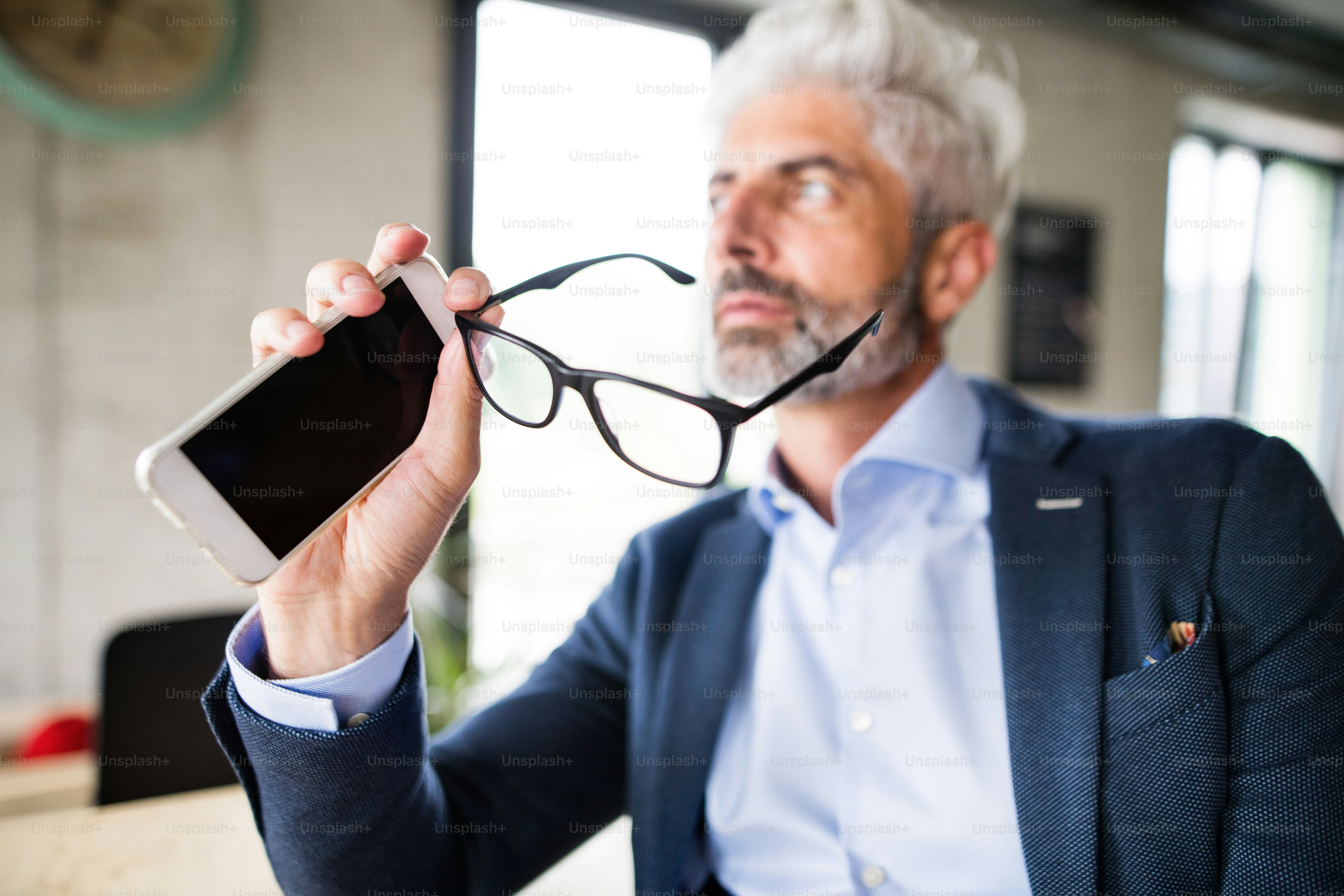 Hübscher, reifer Geschäftsmann mit Smartphone und Brille, der am Schreibtisch im Büro sitzt.