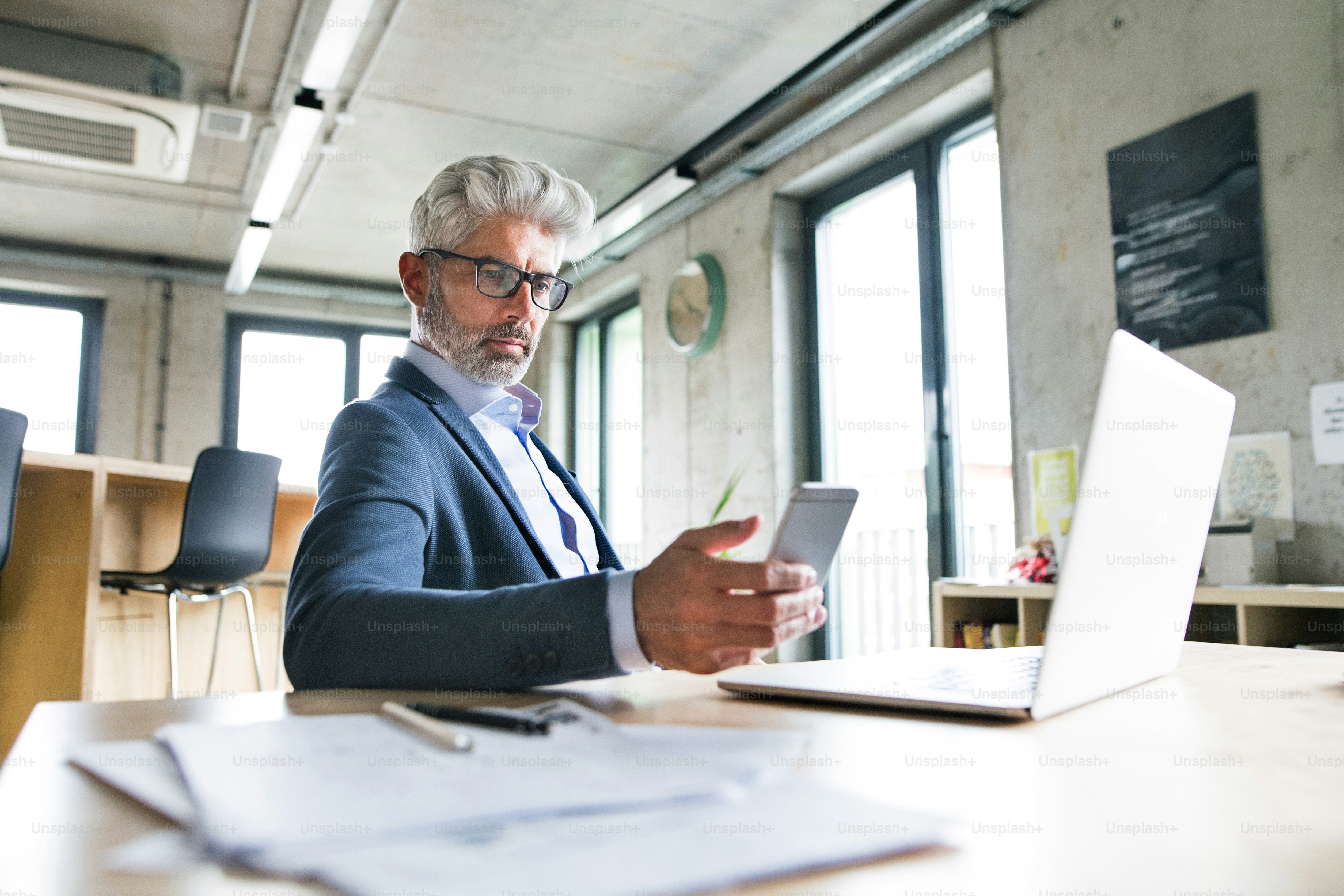 Mature businessman with laptop and smart phone sitting at the desk in the office, texting.