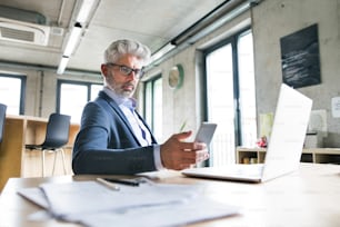 Homme d’affaires mature avec ordinateur portable et téléphone intelligent assis au bureau dans le bureau, textant.