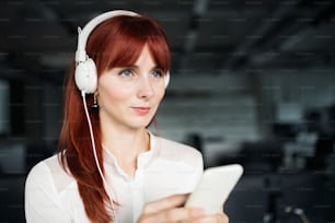 Hermosa joven empresaria en su oficina con grandes auriculares blancos, sosteniendo un teléfono inteligente, escuchando música.