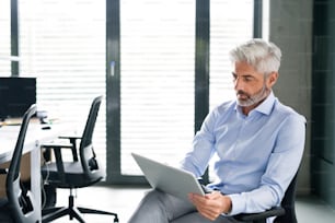 Mature businessman with tablet at the desk in the office, reading documents.