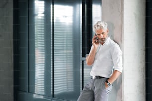 Mature businessman in white shirt in the office standing at the elevator holding smartphone making phone call.