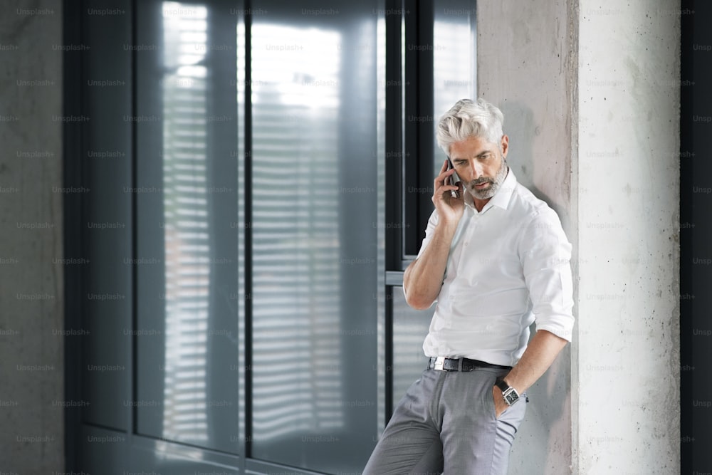 Reifer Geschäftsmann im weißen Hemd im Büro, der am Aufzug steht und ein Smartphone in der Hand hält und telefoniert.