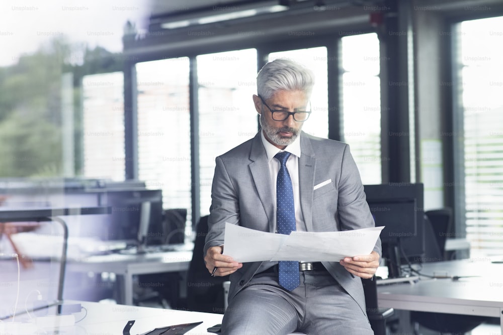 灰色のスーツを着た成熟した実業家がオフィスの机に座って文書を読む。
