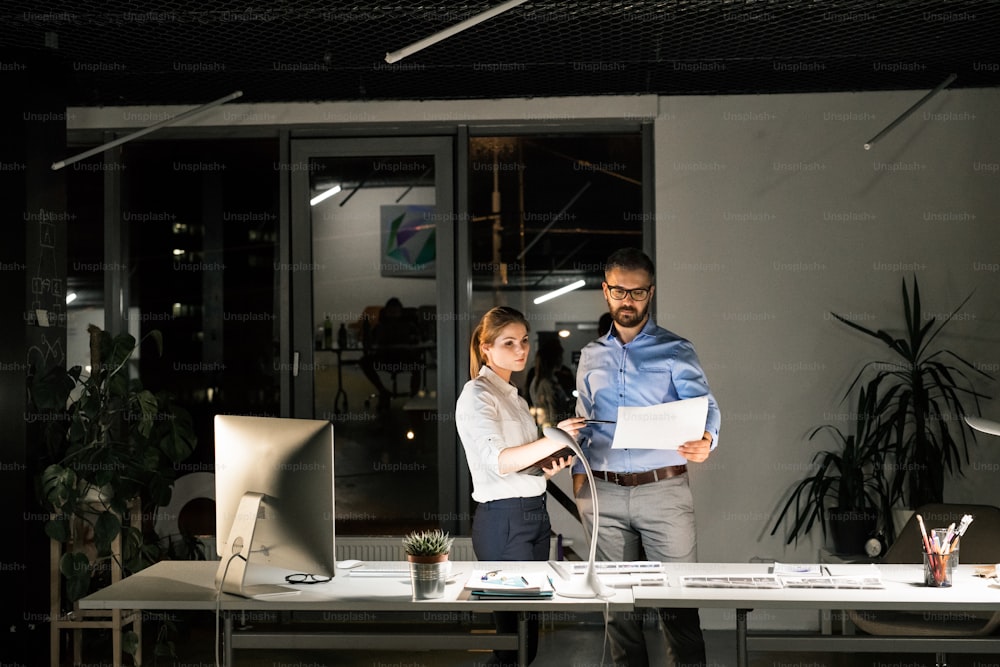 Dos jóvenes empresarios en la oficina por la noche trabajando hasta tarde, discutiendo un proyecto.