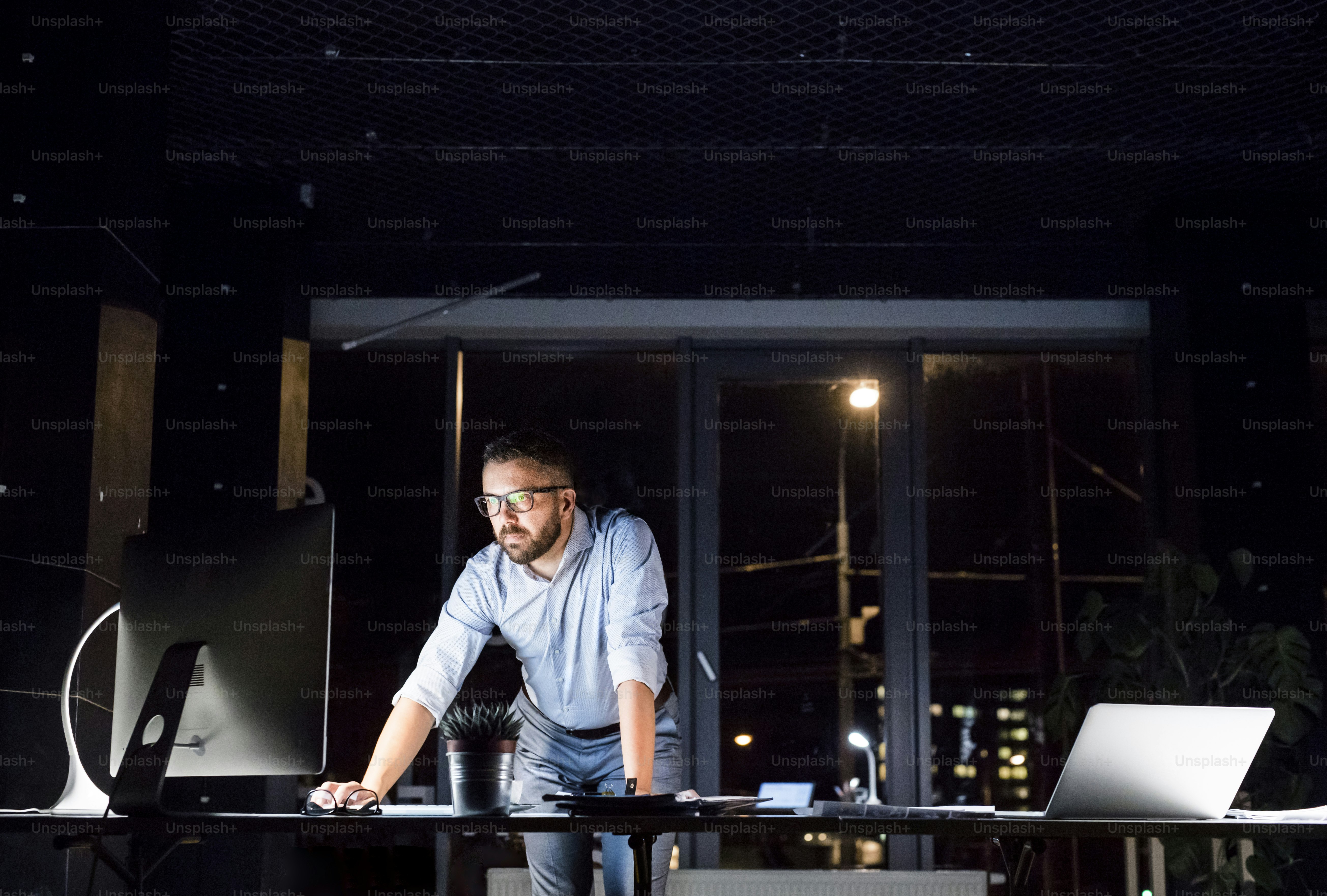 Thoughtful hipster businessman in his office working late at night at desk with computer and laptop.