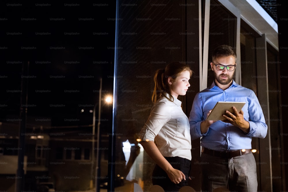 Zwei junge Geschäftsleute mit Tablet im Büro in der Nacht, die spät arbeiten und ein Projekt besprechen.