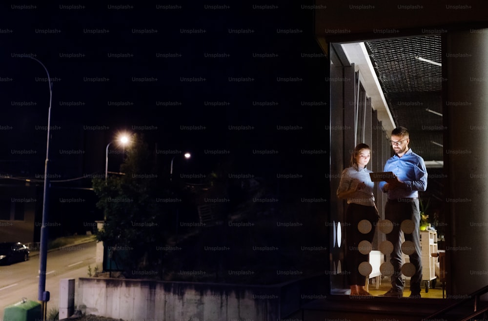 Due giovani imprenditori con tablet in ufficio di notte lavorano fino a tardi, discutendo di un progetto. Girato attraverso il vetro.
