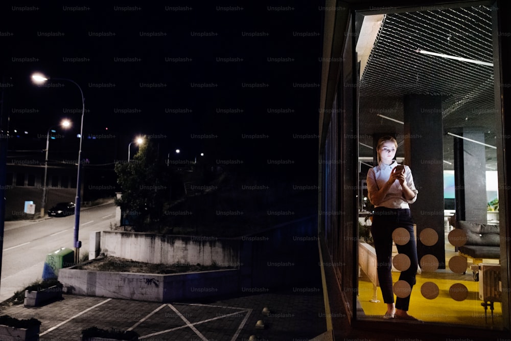 밤늦게까지 일하는 사무실에서 태블릿을 들고 있는 아름다운 젊은 사업가. 유리를 통해 촬영했습니다.