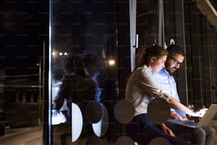 Two young businesspeople in the office at night working late, discussing a project. Shot through the the glass.