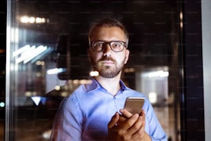 Uomo d'affari hipster riflessivo con smartphone nel suo ufficio che lavora fino a tarda notte.