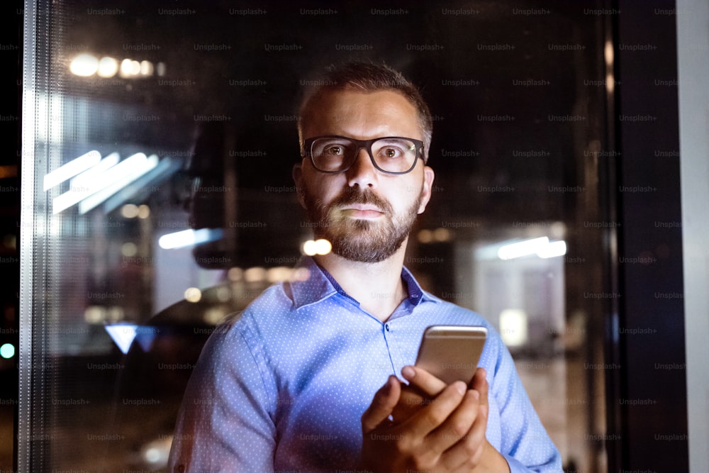 Thoughtful hipster businessman with smart phone in his office working late at night.