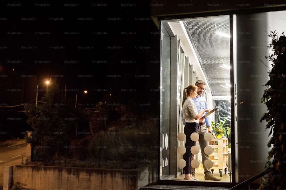Due giovani imprenditori con tablet in ufficio di notte lavorano fino a tardi, discutendo di un progetto. Girato attraverso il vetro.