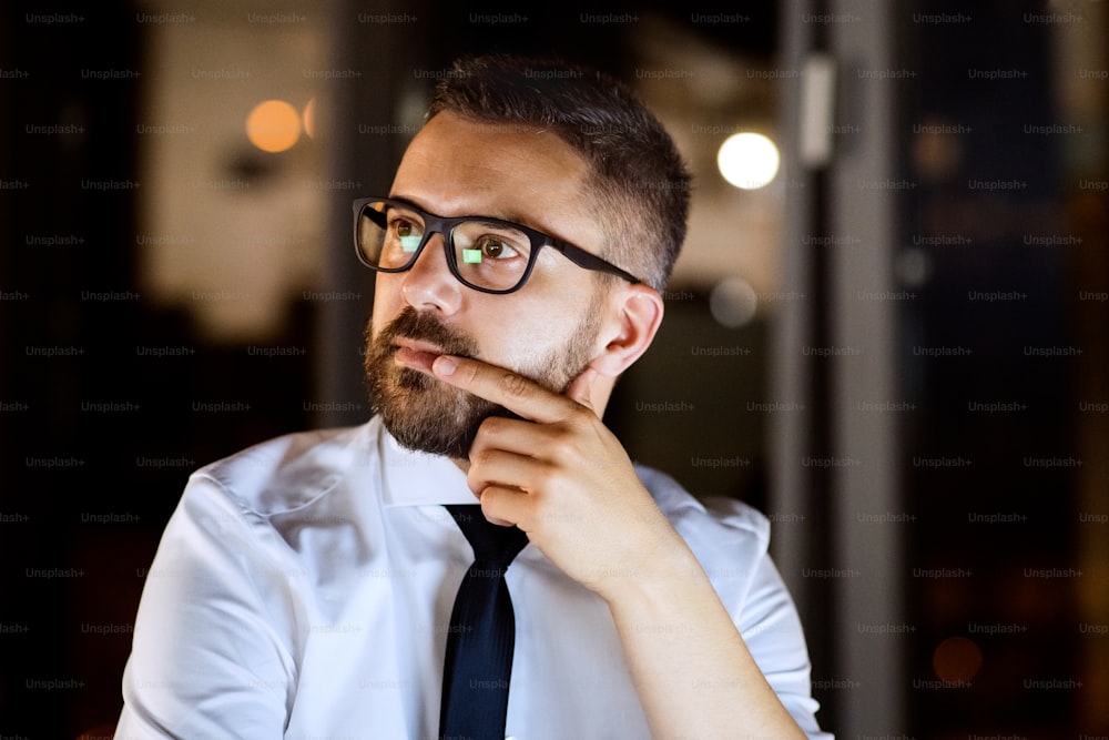 Joven empresario hipster con anteojos negros en su oficina a altas horas de la noche, sentado en el escritorio.