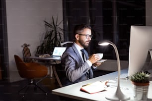 Handsome hipster businessman in his office late at night, sitting at the desk, computer in front of him, drinking coffee.