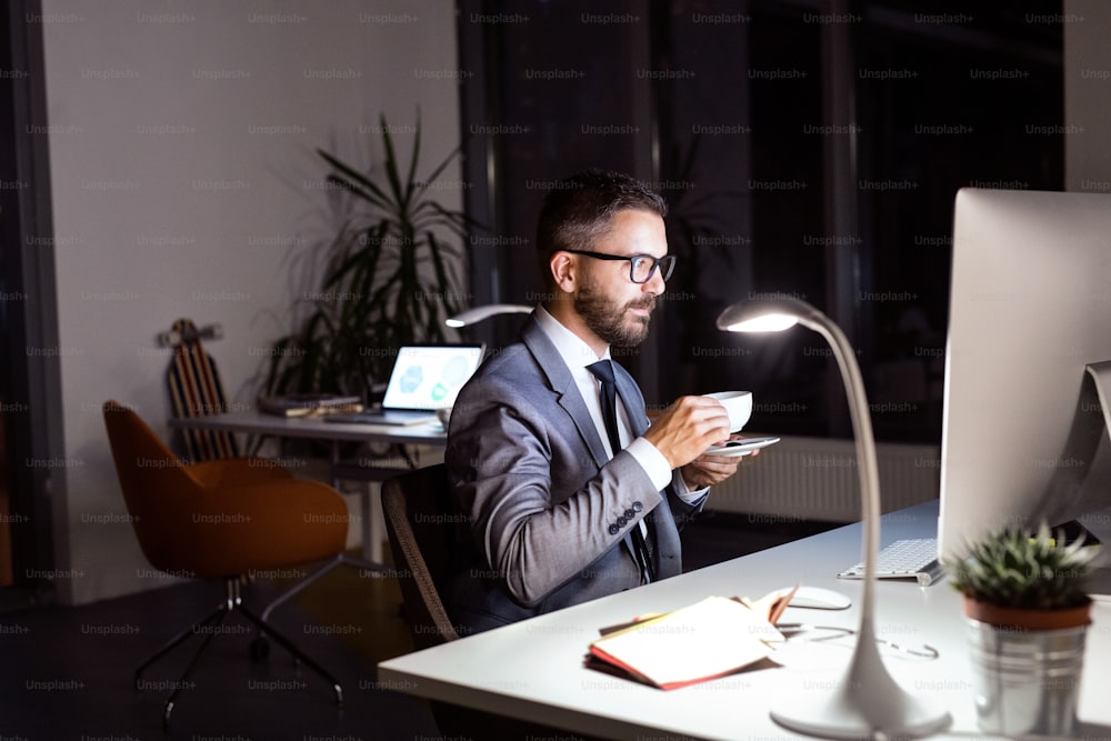 Guapo hombre de negocios hipster en su oficina a altas horas de la noche, sentado en el escritorio, con la computadora frente a él, bebiendo café.