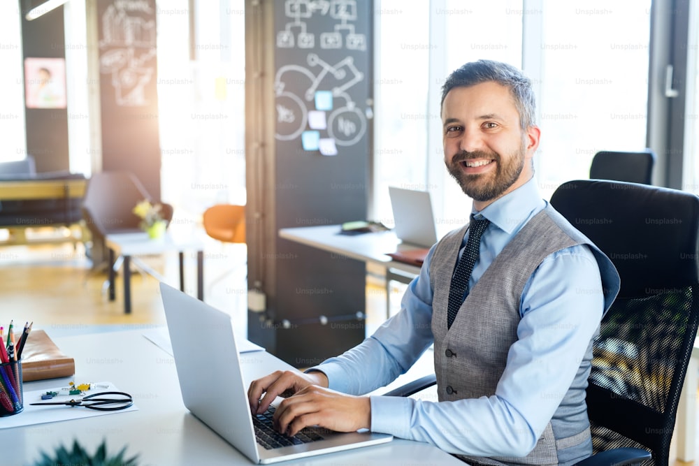 Handsome young businessman in his office, sitting at the desk, laptop in front of him, smiling