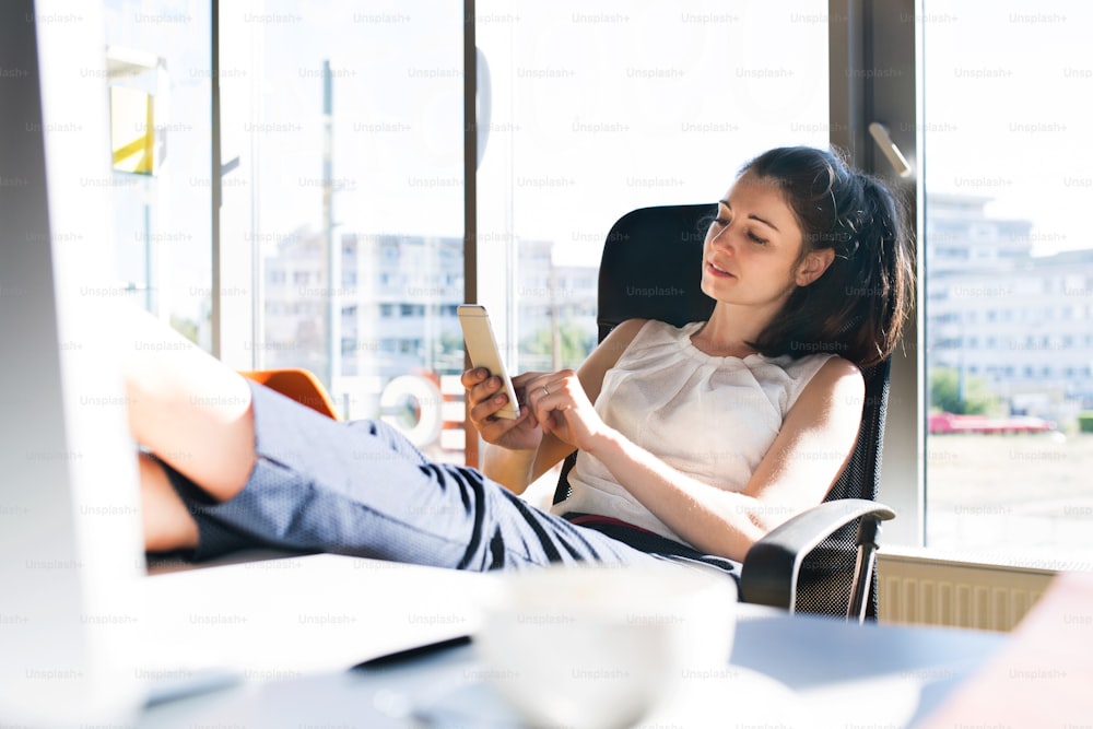 Belle jeune femme d’affaires avec un smartphone dans son bureau assise avec les jambes sur le bureau.