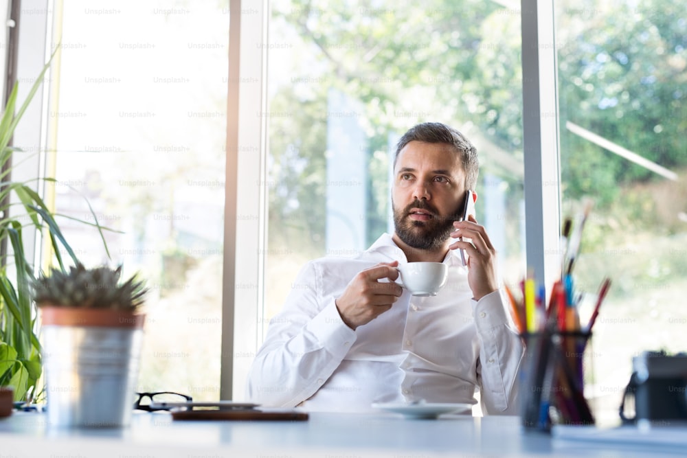 Young hipster businessman working in his office, sitting at the desk, holding coffee and smart phone, making phone call.