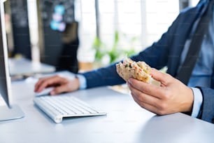 Unrecognizable young businessman in his office, sitting at the desk, computer in front of him, eating his lunch.