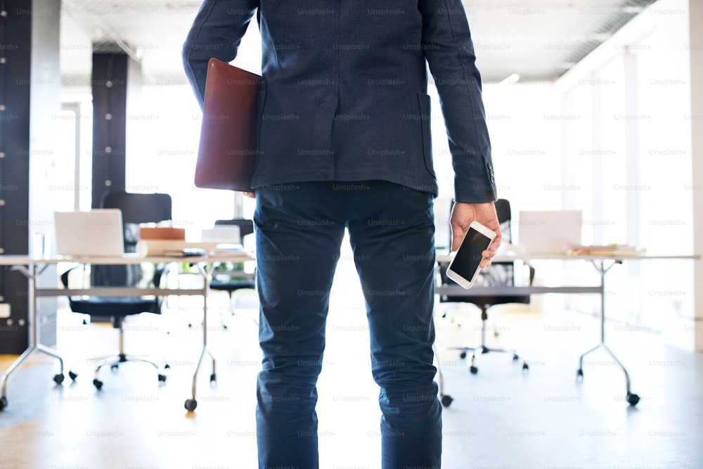 Empresário irreconhecível de pé em seu escritório segurando smartphone e pasta. Vista traseira.