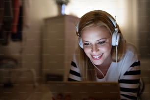 Young beautiful woman lying on the floor, looking at the screen of a tablet late at night, headphones on head.