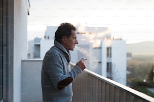 Uomo anziano serio in piedi sul balcone e fumando una sigaretta