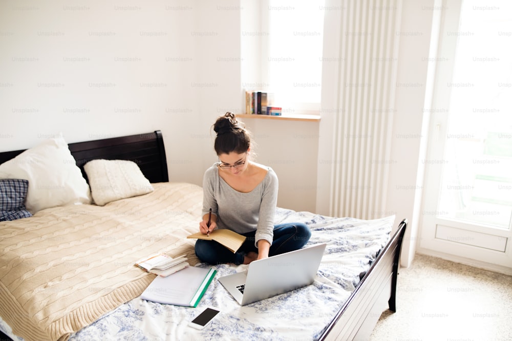 Hermosa joven sentada en la cama, trabajando en una computadora portátil, escribiendo algo en su cuaderno, oficina en casa.