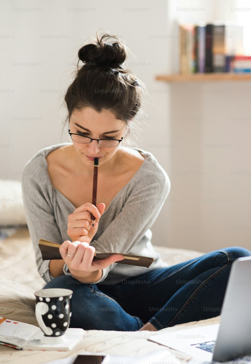 Hermosa joven sentada en la cama, trabajando en una computadora portátil, escribiendo algo en su cuaderno, oficina en casa.