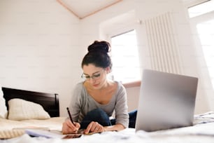 Beautiful young woman sitting on bed, working on laptop, writing something into her notebook, home office.