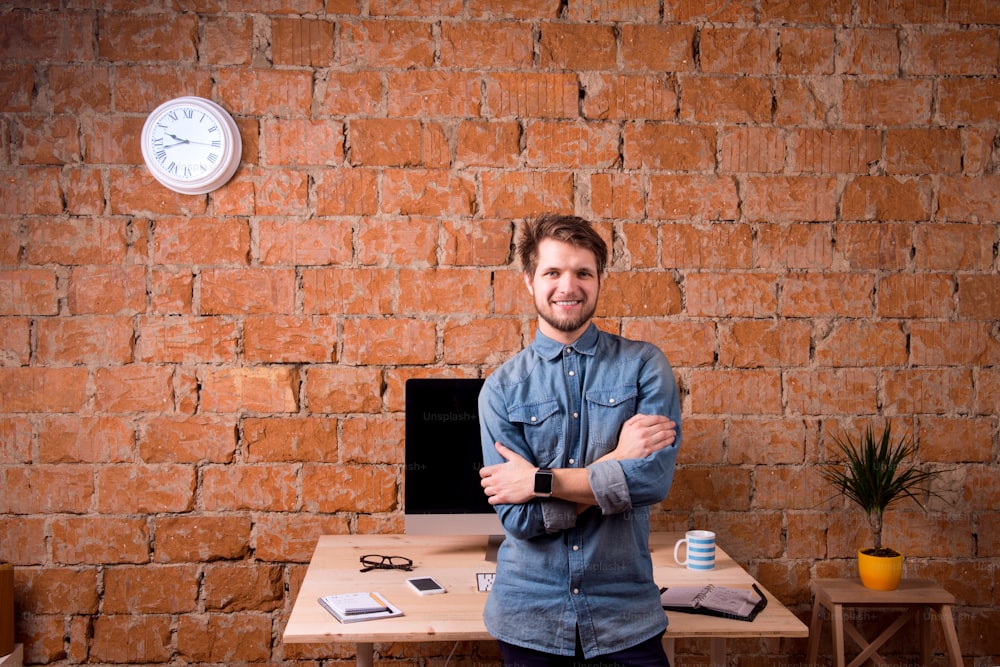 Hipster businessman sitting on office desk, smiling, against brick wall. Smart watch on hand and computer on the table. Coffee cup, personal organizer, smart phone and various office stuff around the workplace.