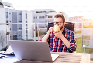 Hipster businessman with laptop in checked shirt sitting on a balcony, holding a cup of coffee, relaxing